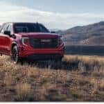 A red 2026 GMC Sierra 1500 pickup truck navigates a grassy off-road trail, set against a scenic backdrop of rolling hills and mountains.
