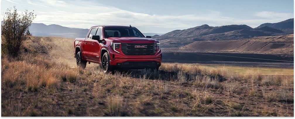 A red 2026 GMC Sierra 1500 pickup truck navigates a grassy off-road trail, set against a scenic backdrop of rolling hills and mountains.