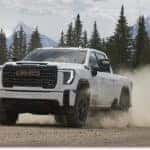 A white 2026 GMC Sierra 2500 HD heavy-duty pickup truck kicks up a large cloud of dust while driving along a dirt road, set against a scenic backdrop of snow-capped mountains and pine trees.