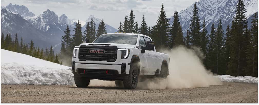 A white 2026 GMC Sierra 2500 heavy-duty pickup truck kicks up a large cloud of dust while driving along a dirt road, set against a scenic backdrop of snow-capped mountains and pine trees.