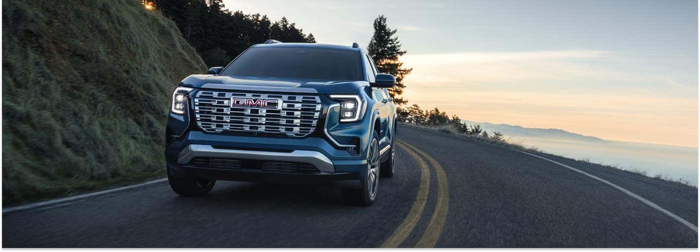 Front three-quarter view of a blue 2026 GMC Terrain SUV driving on a coastal road at sunset.