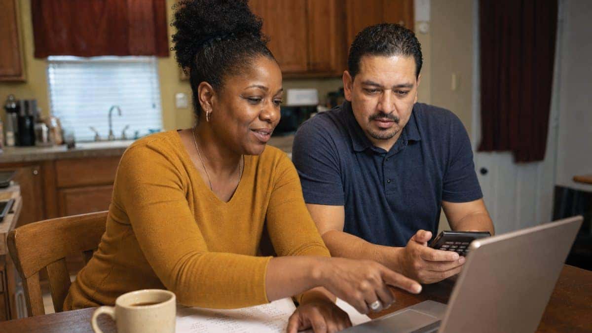 Illustration: a couple at their kitchen table comparing vehicle costs on a laptop with a calculator and handwritten notes