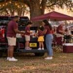 Illustration: a family sets out food on a pickup truck tailgate under pecan trees and string lights while friends carry chairs and cook at a college football tailgate