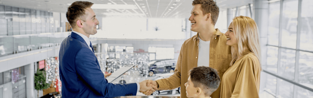 Smiling couple with child selling their vehicle at a dealership, shaking hands with a salesperson