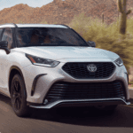 A white Toyota Highlander Hybrid drives down a desert road past cacti and bushes.