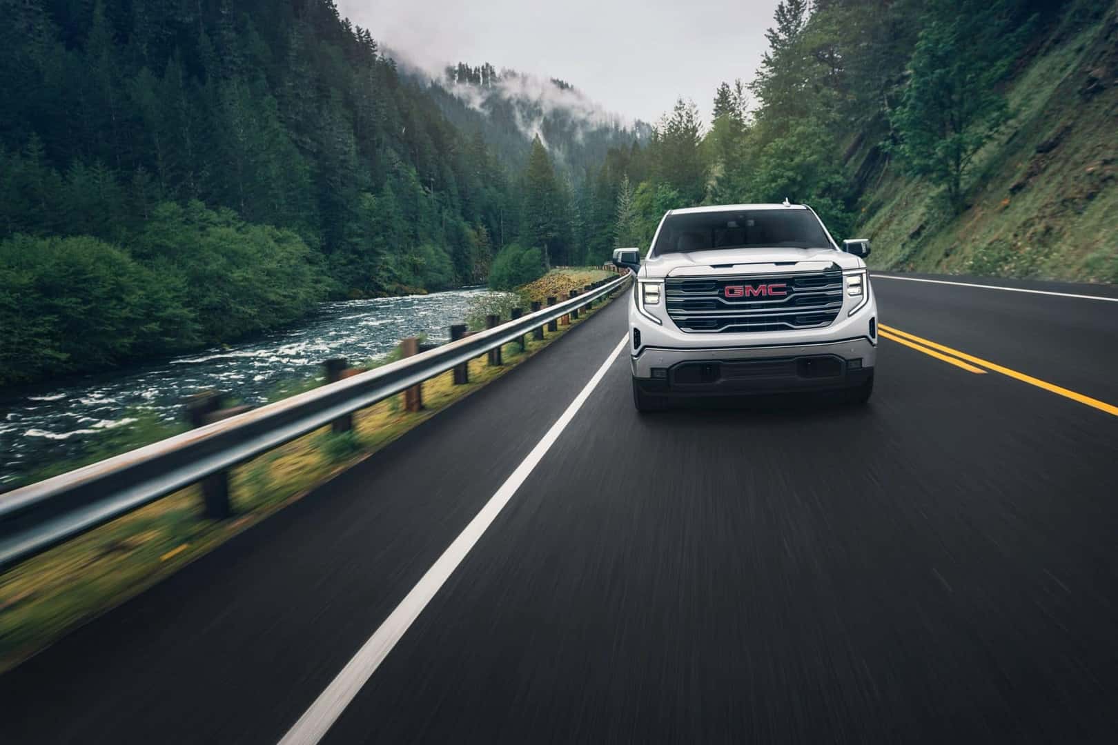 The Confident White 2024 GMC Sierra Strolling On A Road, Near The Woodland Hillside