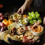 Close-up of a woman serving a charcuterie cheese and meat platter with fruits. Female hands placing mixed fruits with cheese and meat on wooden board.