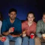 Multi-ethnic group of friends watching movies at home while eating snacks and popcorn sitting on big sofa in dark room