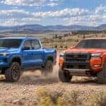 Blue 2026 Chevrolet Colorado ZR2 and orange 2026 Nissan Frontier PRO-4X driving on a dirt road in open country