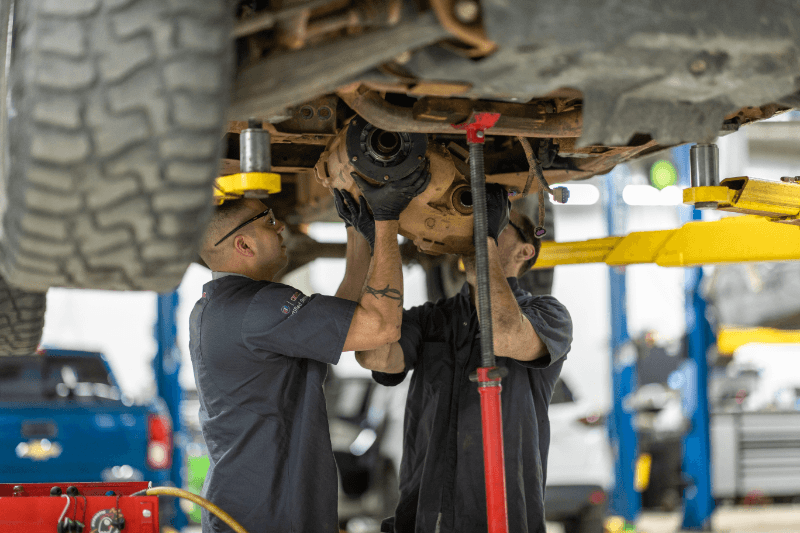 Two Weld County Garage technicians diagnosing a driveline problem under a lifted truck