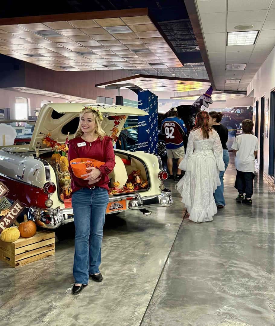 A Weld County Garage team member holds a bowl of candy beside a classic car with a pumpkin patch scene in its trunk as costumed trick-or-treaters walk past.