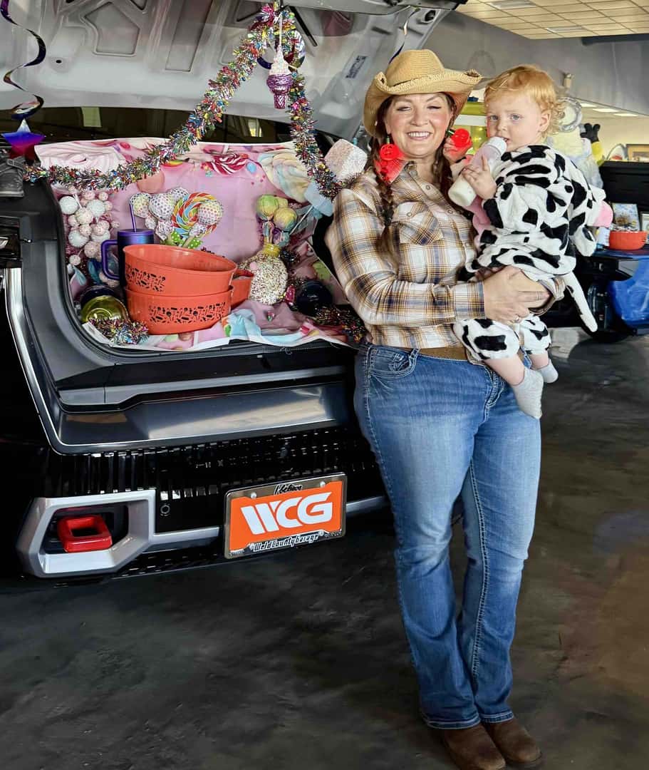 A woman in a cowgirl costume holds a toddler in a cow costume next to a GMC with a candy-themed display in its trunk at Weld County Garage.
