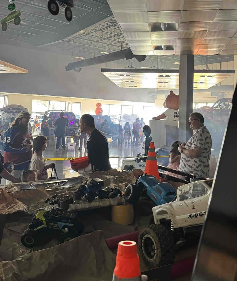 Families trick-or-treating in the fog-filled Weld County Garage showroom during a past Trunk or Treat, with a table of RC trucks in the foreground.